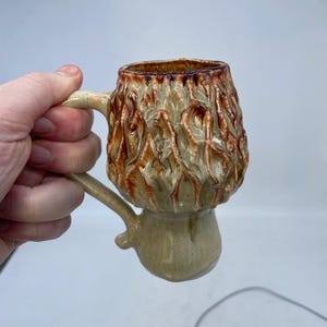 Peut inclure: Une tasse en céramique faite à la main avec un design texturé et organique. La tasse présente une base beige avec une partie supérieure texturée, marron et beige. Elle a une anse et un bord marron foncé. La tasse est conçue pour les boissons.