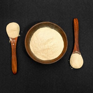 May include: A top-down shot features a small, round, brown bowl filled with a light beige powder. Two wooden spoons, each holding a similar amount of powder, flank the bowl. The background is a solid black surface.