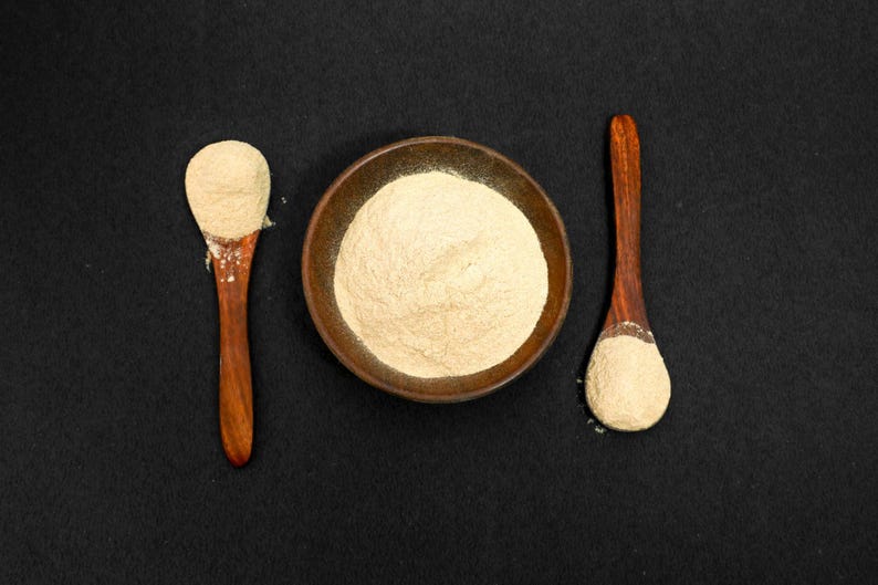 May include: A top-down shot features a small, round, brown bowl filled with a light beige powder. Two wooden spoons, each holding a similar amount of powder, flank the bowl. The background is a solid black surface.