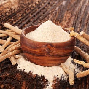 May include: A wooden bowl filled with light brown powder, surrounded by tan-colored sticks, sits on a dark wooden surface. The powder spills out of the bowl, and the sticks are scattered around it. The image suggests a natural health product.
