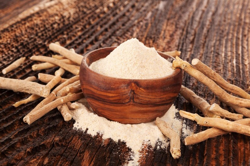 May include: A wooden bowl filled with light brown powder, surrounded by tan-colored sticks, sits on a dark wooden surface. The powder spills out of the bowl, and the sticks are scattered around it. The image suggests a natural health product.