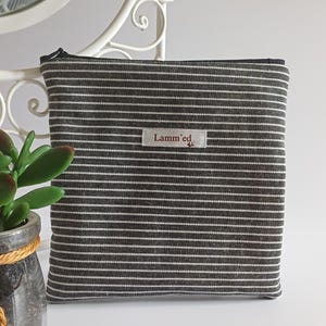 Könnte beinhalten: Eine rechteckige Tasche mit schwarz-weißem Streifenmuster. Die Tasche hat einen schwarzen Reißverschluss und ein kleines Etikett mit der Aufschrift "Lamm'ed". Die Tasche liegt auf einer weißen Oberfläche.
