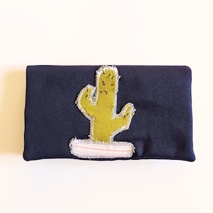 Può includere: Pochette blu navy con un'applicazione a cactus giallo. Il cactus è cucito con un filo strutturato verde chiaro e poggia su una base a righe rosa e bianche. La pochette è rettangolare e sembra essere fatta di tessuto.