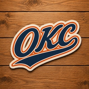 Könnte beinhalten: Ein cremefarbener Aufkleber mit den Buchstaben "OKC" in einer stilisierten Schriftart. Die marineblauen Buchstaben haben eine orangefarbene Umrandung auf cremefarbenem Hintergrund. Der Aufkleber liegt auf einer Holzoberfläche.