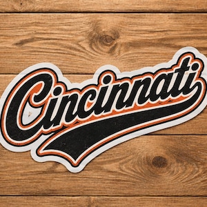 May include: A die-cut sticker with the word "Cincinnati" in a script font. The text is black with an orange outline and a white border. The sticker is on a wooden background.
