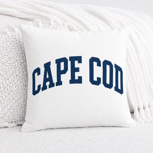 May include: A square white throw pillow featuring the text CAPE COD printed in a bold, dark blue, arched collegiate-style font across the center. The pillow has a smooth, solid white fabric surface and clean, straight edges on all four sides.