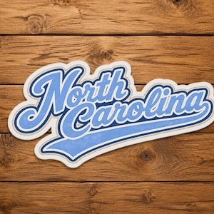 May include: A sticker with the words "North Carolina" in a stylized, light blue script with a white and dark blue outline. The sticker is on a wooden background.