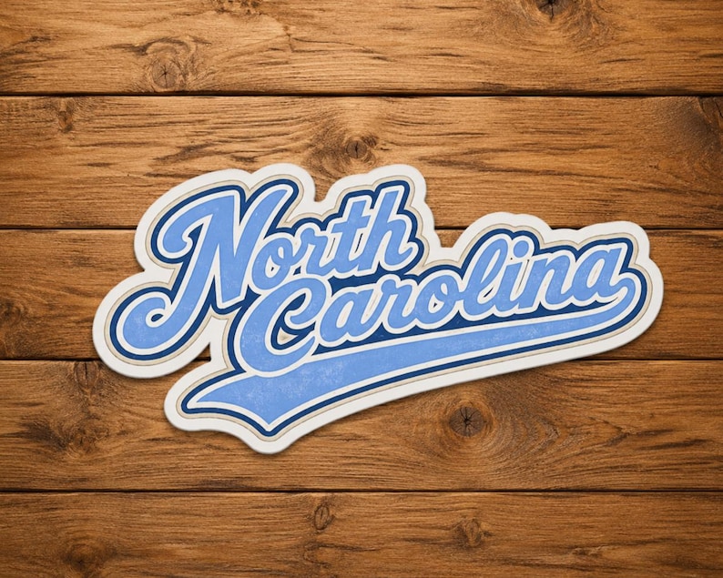 May include: A sticker with the words "North Carolina" in a stylized, light blue script with a white and dark blue outline. The sticker is on a wooden background.