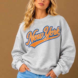 May include: A light gray crewneck sweatshirt featuring the text New York in a bold orange script font with a dark blue outline. The long-sleeved garment has a ribbed collar, cuffs, and hem, providing a classic fit with a casual aesthetic.