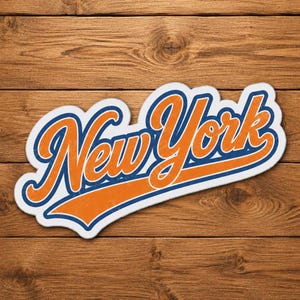 Könnte beinhalten: Ein weißer Aufkleber mit dem Schriftzug "New York" in orangefarbener Schrift mit blauem Rand und blauem Streifen. Der Aufkleber befindet sich auf einem Holzgrund.