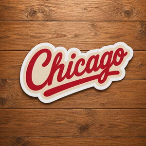 Puede incluir: Una pegatina con la palabra "Chicago" en cursiva roja, con un contorno crema y una línea roja. La pegatina está sobre un fondo de madera.