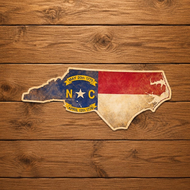 May include: A distressed sticker in the shape of North Carolina, featuring the state flag design. The flag is divided into sections of blue, red, and white, with the letters "N" and "C" and a star. The sticker is on a wooden background.
