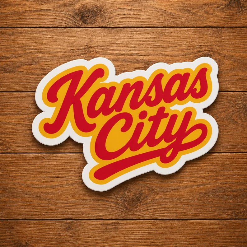 May include: A sticker with the words "Kansas City" in red script lettering with a yellow outline and a white border. The sticker is set against a wooden background.