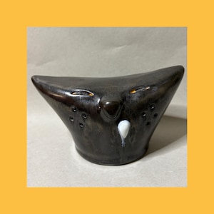 May include: A dark brown ceramic figurine shaped like a stylized cat head with pointed ears. The piece features narrow, orange-accented eyes, a small nose, and a single white teardrop-shaped mark below the nose, with small circular indentations.