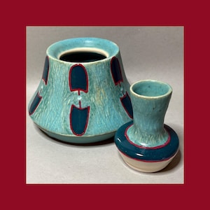 May include: A turquoise ceramic vessel with a wide, tapered base and a smaller, matching turquoise ceramic bottle. Both pieces feature a textured surface, dark blue accents, and thin red outlines around the decorative shapes and rims.