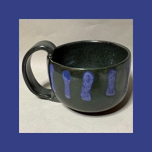 May include: A dark green ceramic mug with a glossy finish and a large, curved handle. The exterior features vertical, textured blue drip patterns spaced evenly around the body, creating a distinct contrast against the deep green base color.