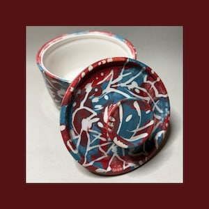 May include: A round ceramic container with a matching lid, featuring a vibrant splatter pattern in shades of red, blue, and white. The interior of the container is smooth and white, while the exterior and lid display a dynamic, multi-colored design.