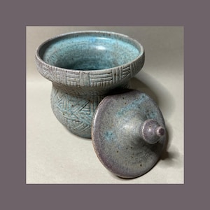 May include: A ceramic jar with a lid featuring a textured, geometric pattern. The jar has a wide, open mouth and a rounded body, finished in a mottled glaze of turquoise, blue, and grey tones. The matching lid has a small, pointed handle.