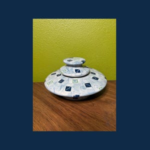 May include: A ceramic trinket box with a lid, featuring a light blue base and lid with a pattern of blue and green squares. The box is round with a small, stacked lid on top. The background is a wooden surface and a green wall.