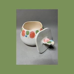 May include: A white ceramic jar with a lid, decorated with colorful, raised oval shapes in red, green, and blue. The lid is off to the side, revealing the interior of the jar. The jar has a rounded shape and a smooth finish.