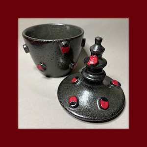 May include: A dark gray ceramic lidded jar with a speckled finish. The jar and its conical lid are decorated with small, raised red and black oval accents. The lid features a tiered, pointed finial handle at the top, and the jar has a wide opening.
