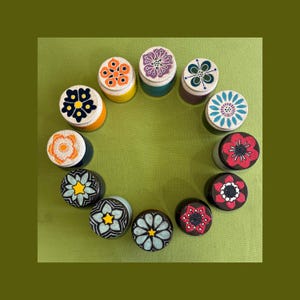 May include: Twelve small cylindrical containers arranged in a circle. Each container features a unique, hand-painted floral design on its lid, showcasing a variety of colors including orange, purple, green, blue, red, and black patterns.