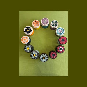 May include: A collection of twelve small, cylindrical containers arranged in a circle. Each lid features a unique, hand-painted floral or geometric pattern in various colors including red, blue, yellow, orange, purple, and black.