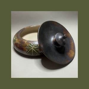 May include: A round ceramic lidded jar with a dark brown and tan mottled glaze. The exterior features embossed green leaf motifs with white veining. The lid is circular, dark brown, and includes a small central knob for easy handling.