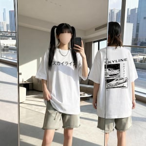 Könnte beinhalten: Weißes T-Shirt mit japanischem Text auf der Vorderseite und einer Autografik auf der Rückseite. Das Shirt wird mit olivgrünen Shorts getragen. Auf der Rückseite des Shirts steht "SKYLINE" mit japanischem Text und einer Autografik.