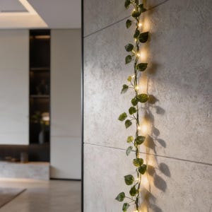 Puede incluir: Una guirnalda decorativa de luces LED con hojas verdes artificiales, que desciende verticalmente por una pared gris texturizada. Las luces blancas cálidas crean un brillo suave, realzando el aspecto natural del follaje artificial.