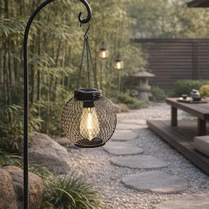 Könnte beinhalten: Eine schwarze Metall-Außenlaterne mit Drahtgitter-Design, die an einem gebogenen Pfosten hängt. Die Laterne hat ein warmes, leuchtendes Licht im Inneren. Der Hintergrund zeigt einen Garten mit Steinwegen und Bambus.