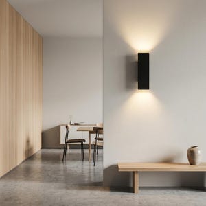 Puede incluir: Un interior moderno con una lámpara de pared rectangular negra que emite luz cálida hacia arriba y hacia abajo. La lámpara está montada en una pared gris claro. La habitación también incluye un banco de madera, un jarrón y un vistazo a un comedor.