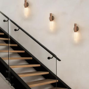 Puede incluir: Tres apliques de pared con respaldo de madera y una pantalla cilíndrica de vidrio esmerilado están montados en una pared blanca. Los apliques están cerca de una escalera con escalones de madera y una barandilla de metal negro.