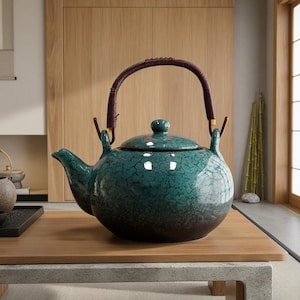 Può includere: Una teiera in ceramica turchese con base marrone scuro e manico intrecciato. La teiera ha un coperchio e un beccuccio. Il manico è marrone scuro e la teiera è appoggiata su una superficie di legno.