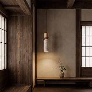 Puede incluir: Una moderna lámpara colgante con una pantalla cilíndrica texturizada y una parte superior de madera. La luz cuelga de un cable negro en una habitación con paredes de madera y un banco. Un pequeño jarrón con ramas está sobre el banco.