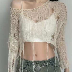 Peut inclure: Haut en tricot beige à mailles ouvertes, avec des manches longues et une coupe courte. Le haut a un aspect usé avec des trous intentionnels et des fils pendants. Un caraco blanc est visible en dessous. Le haut est associé à un jean délavé.
