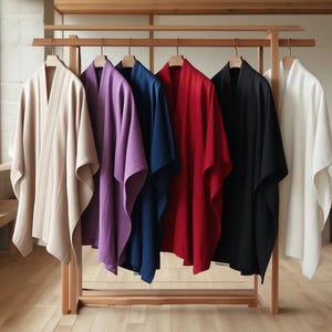 Puede incluir: Un perchero de madera exhibe seis prendas drapeadas en varios colores: beige, morado, azul marino, rojo, negro y blanco. Las prendas tienen un diseño sencillo y fluido, en un espacio minimalista y bien iluminado.