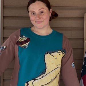 May include: A long-sleeve sweatshirt featuring a teal center panel and brown sleeves. The front displays a woven tapestry-style graphic of a yellow bear and a spinning top. The hem and cuffs are finished with a multi-colored fringe trim detail.