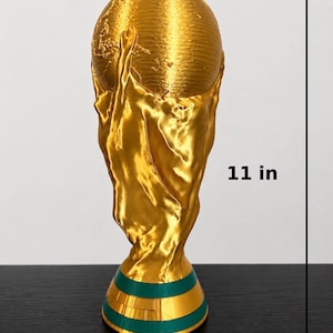 May include: A gold-coloured trophy, approximately 28 cm tall, featuring a globe held by two stylised hands. The base has green and gold bands. The trophy is a representation of a world championship.