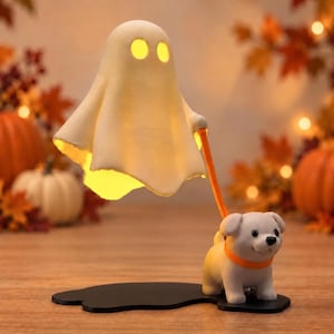 May include: A decorative lamp featuring a white ghost figure with glowing yellow eyes and a mouth, connected by an orange leash to a small white puppy figurine. The entire assembly is mounted on a flat black base with a smooth, rounded shape.