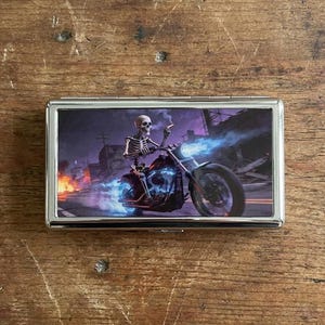 May include: A silver metal cigarette case featuring a skeleton riding a motorcycle with blue flames. The background shows a post-apocalyptic scene with burning buildings and a purple sky. The case is rectangular.