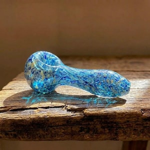 May include: A blue glass pipe with a speckled pattern of light blue and gold. The pipe is resting on a weathered wooden surface, with the light reflecting off the glass.