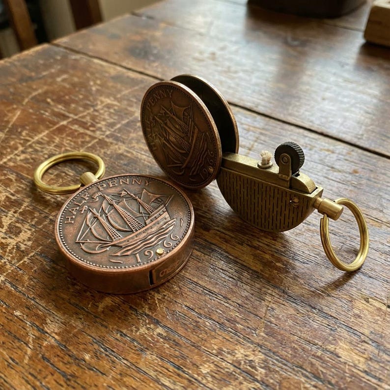 May include: A vintage brass lighter with a Half Penny coin design. The lighter is open, revealing the wick and flint wheel. A matching coin is next to the lighter, and a brass ring is attached. The lighter and coin are on a wooden surface.