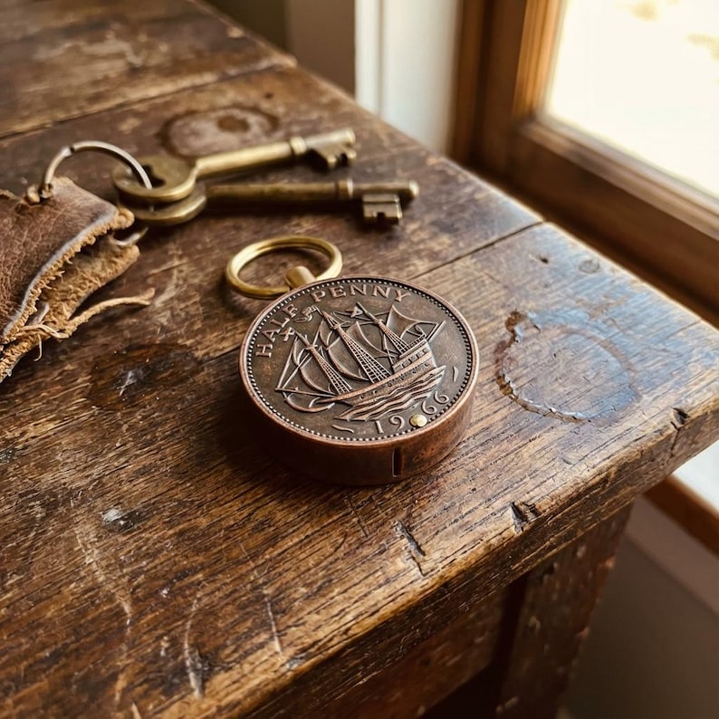 May include: A vintage Half Penny coin from 1966, featuring a sailing ship, is set into a small, round, copper-colored container. The coin is next to a leather key holder, two brass keys, and a key ring, all resting on a weathered wooden surface.