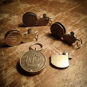 May include: A collection of vintage brass lighters and keychains, some featuring a ship design. The lighters have a classic design, and the keychains are made from coins. The items are arranged on a wooden surface.