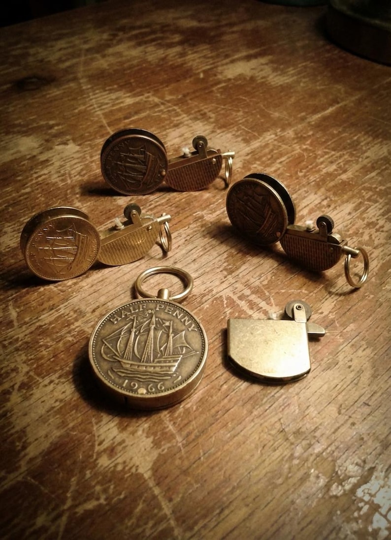 May include: A collection of vintage brass lighters and keychains, some featuring a ship design. The lighters have a classic design, and the keychains are made from coins. The items are arranged on a wooden surface.