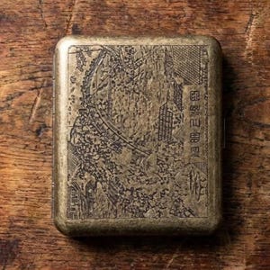 May include: A rectangular brass-toned metal cigarette case featuring an intricate etched landscape scene on the front cover. The design includes detailed depictions of traditional architecture, trees, and foliage, with vertical text characters on the right.