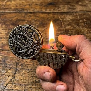Vintage-style brass coin lighter inspired by old British half penny designs. The lid features an engraved ship and the inscription “Half Penny 1966,” giving it a classic maritime look. Made from durable metal with an aged brass finish, it uses a traditional wick and flint ignition system. The detailed coin design and textured body create a unique retro collectible with a strong old-world aesthetic.