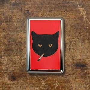 Może przedstawiać: Srebrne etui na papierosy z czerwonym tłem. Przedstawia czarnego kota z żółtymi oczami i papierosem w ustach. Etui ma prostokątny kształt.