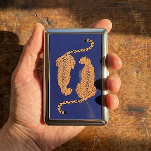 Op de afbeelding: Een rechthoekige, zilverkleurige etui met een marineblauwe oppervlakte met twee cheeta's met oranje en zwarte vlekken. De cheeta's kijken elkaar aan. De etui wordt in een hand gehouden.